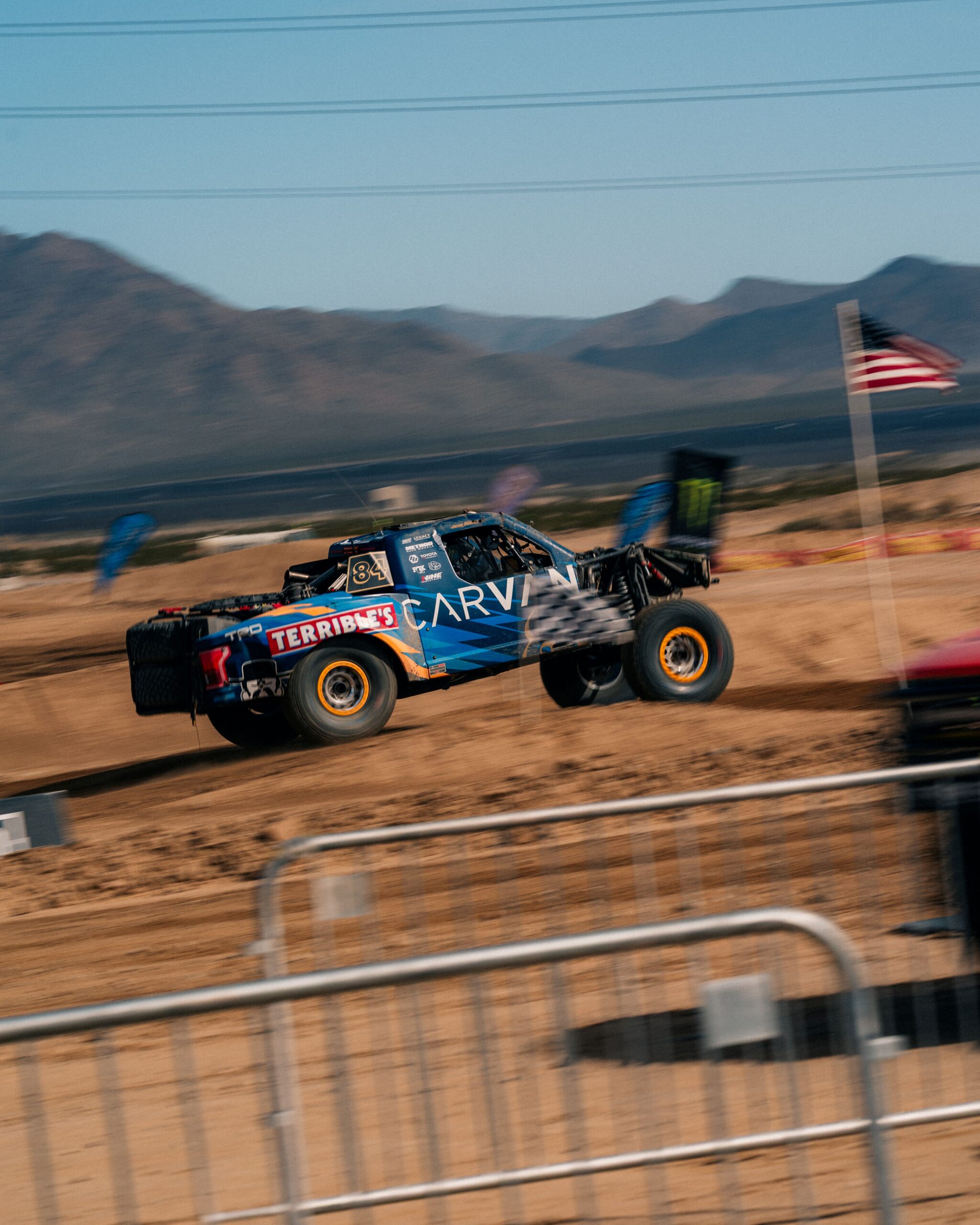 Carvana-sponsored No. 84 Terrible Herbst Motorsports off-road truck racing at speed at the BFGoodrich Mint 400 in Nevada, with motion blur and an American flag nearby.