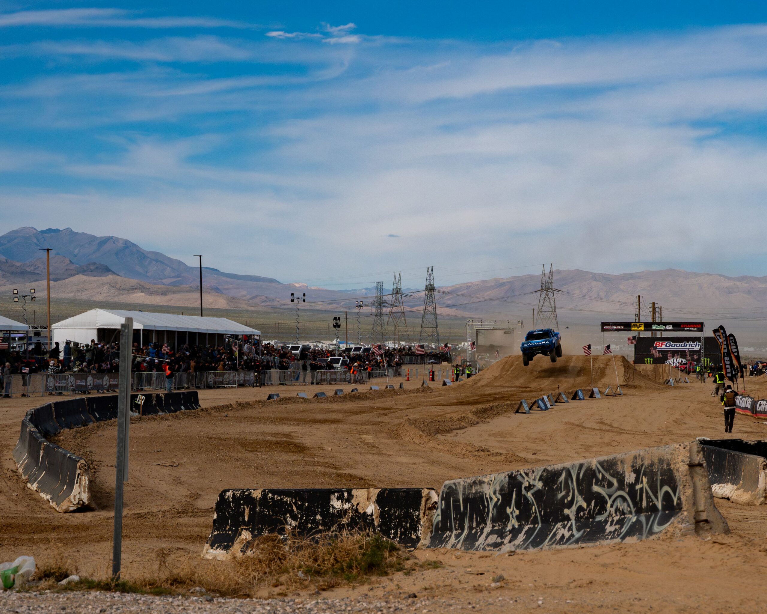Carvana-sponsored off-road truck launching off a jump at the BFGoodrich Mint 400 race course in Nevada with spectators and event signage in the background.