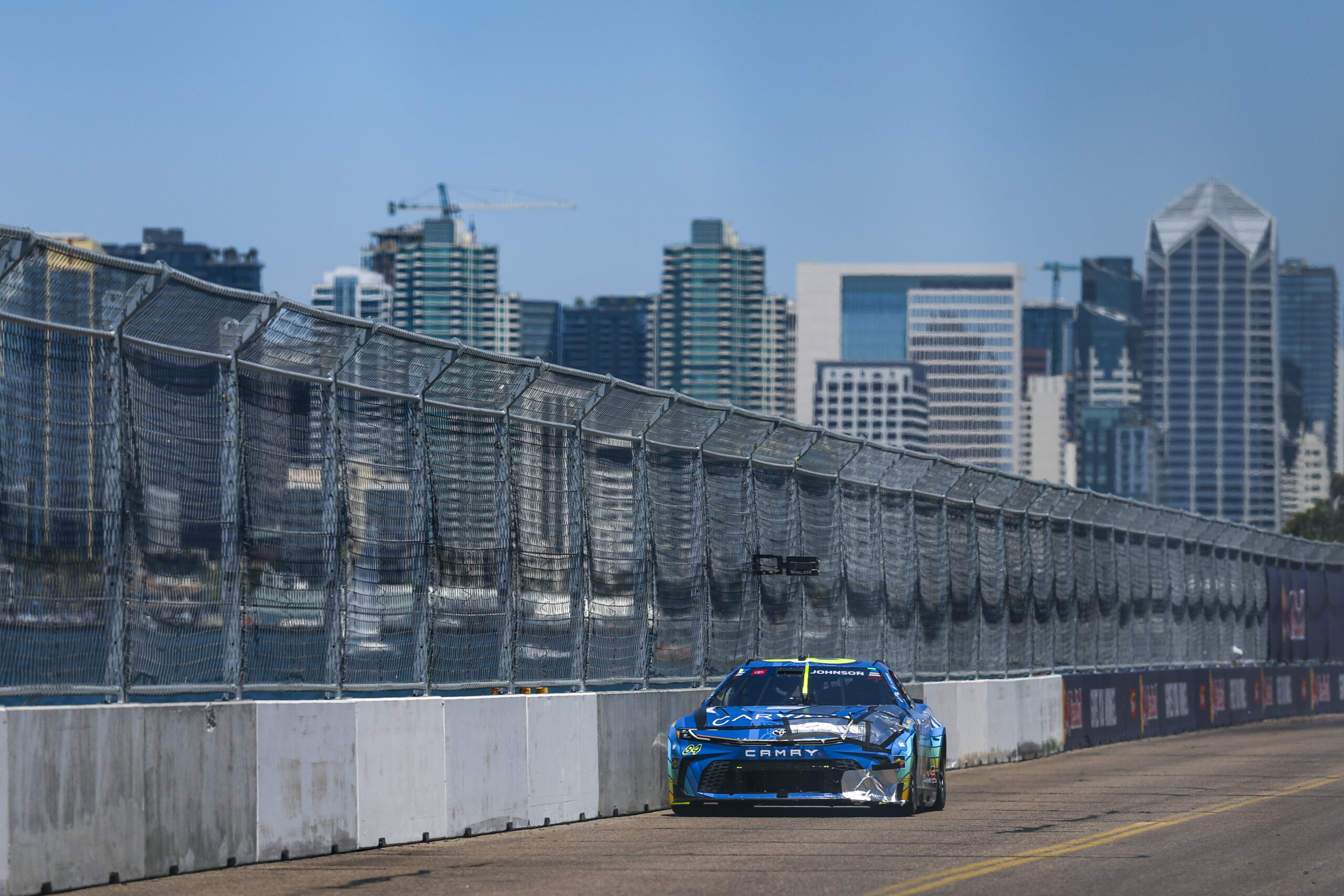 The No. 84 Carvana Toyota Camry XSE races along a straightaway at Naval Base Coronado with the downtown San Diego skyline visible across the bay in the background.