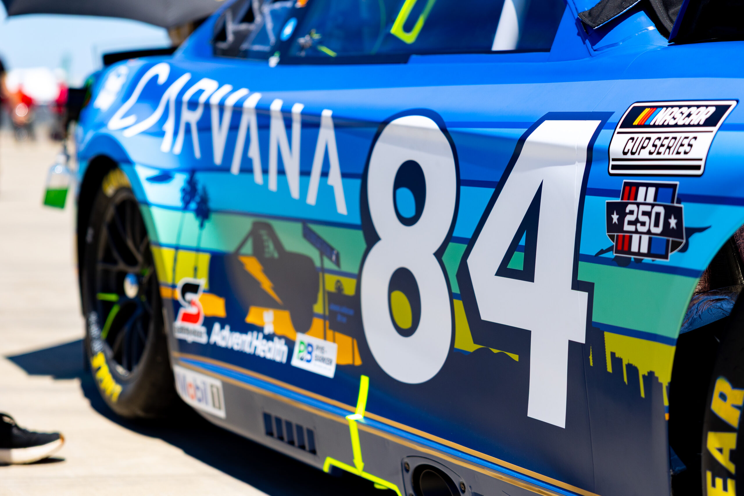 A close-up of the driver's side of the No. 84 Carvana Toyota Camry XSE in the garage at Naval Base Coronado, showing the custom San Diego paint scheme featuring a layered cityscape and landscape design in shades of blue, teal, green, and gold.