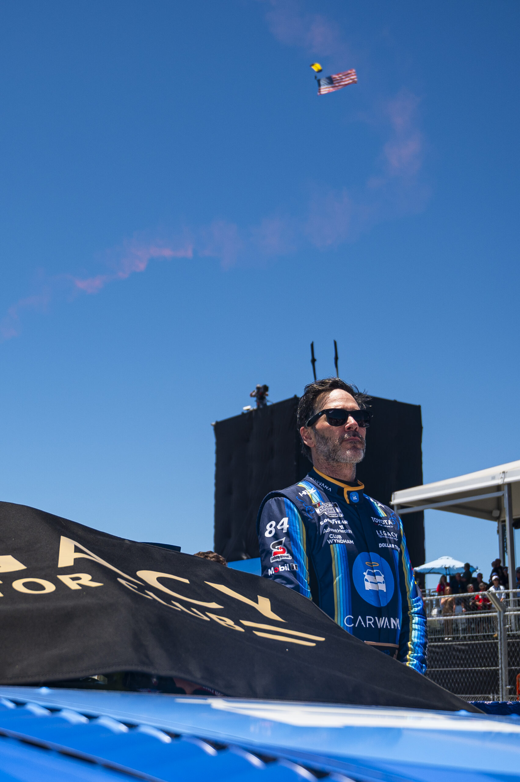 Jimmie Johnson stands on pit road in his No. 84 Carvana Legacy Motor Club firesuit during pre-race ceremonies at Naval Base Coronado, with a skydiver carrying an American flag descending in the background.